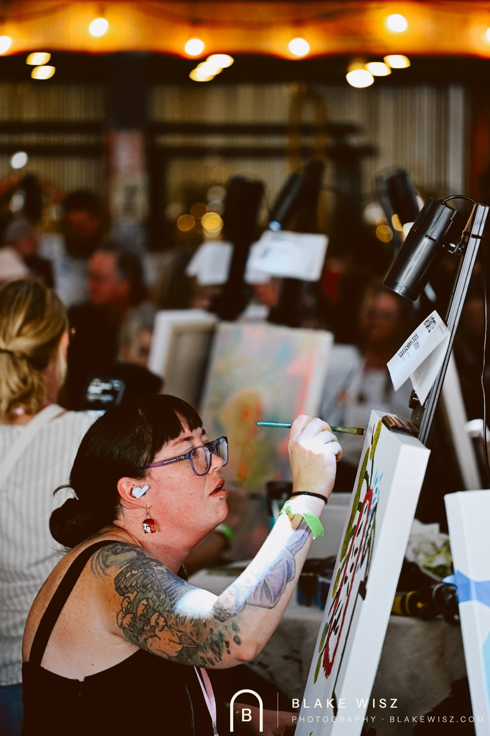 Tattooed artist in glasses painting at her easel under string lights at Art Battle Ormond Beach