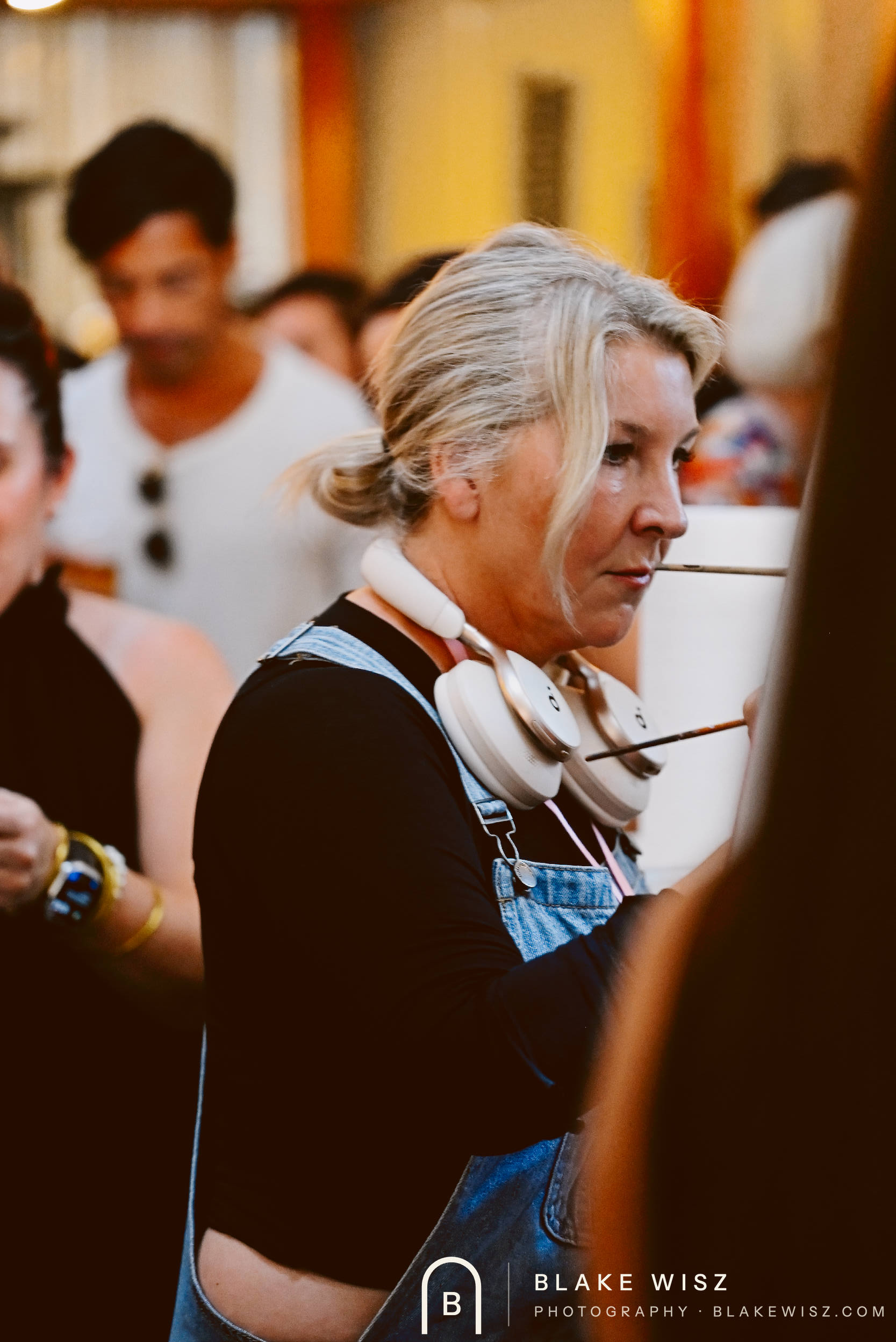 Art Battle champion Michelle A Delisle at her easel in denim overalls, brush between her teeth