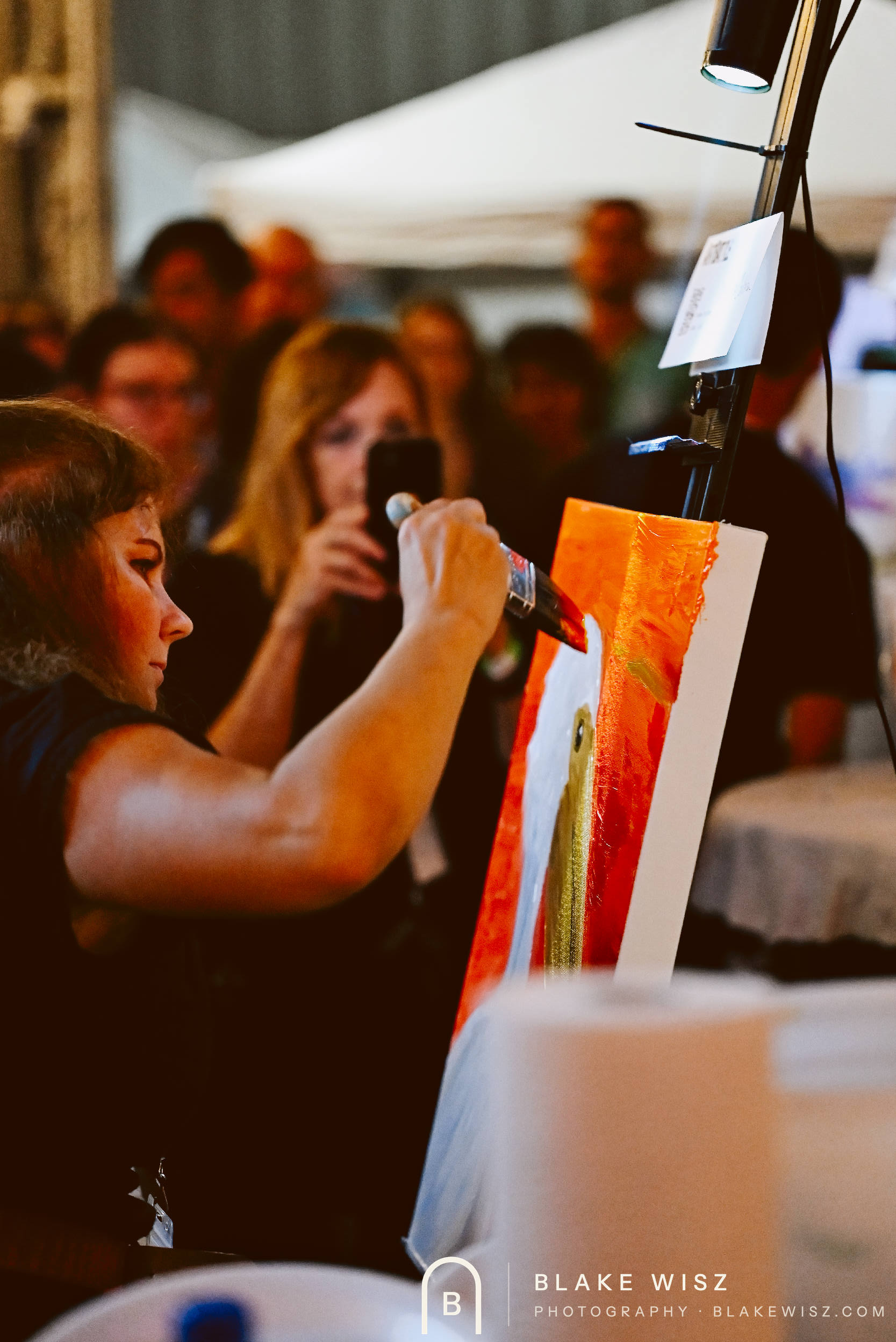 Artist blocking in a white bird on a red canvas as a spectator films the round
