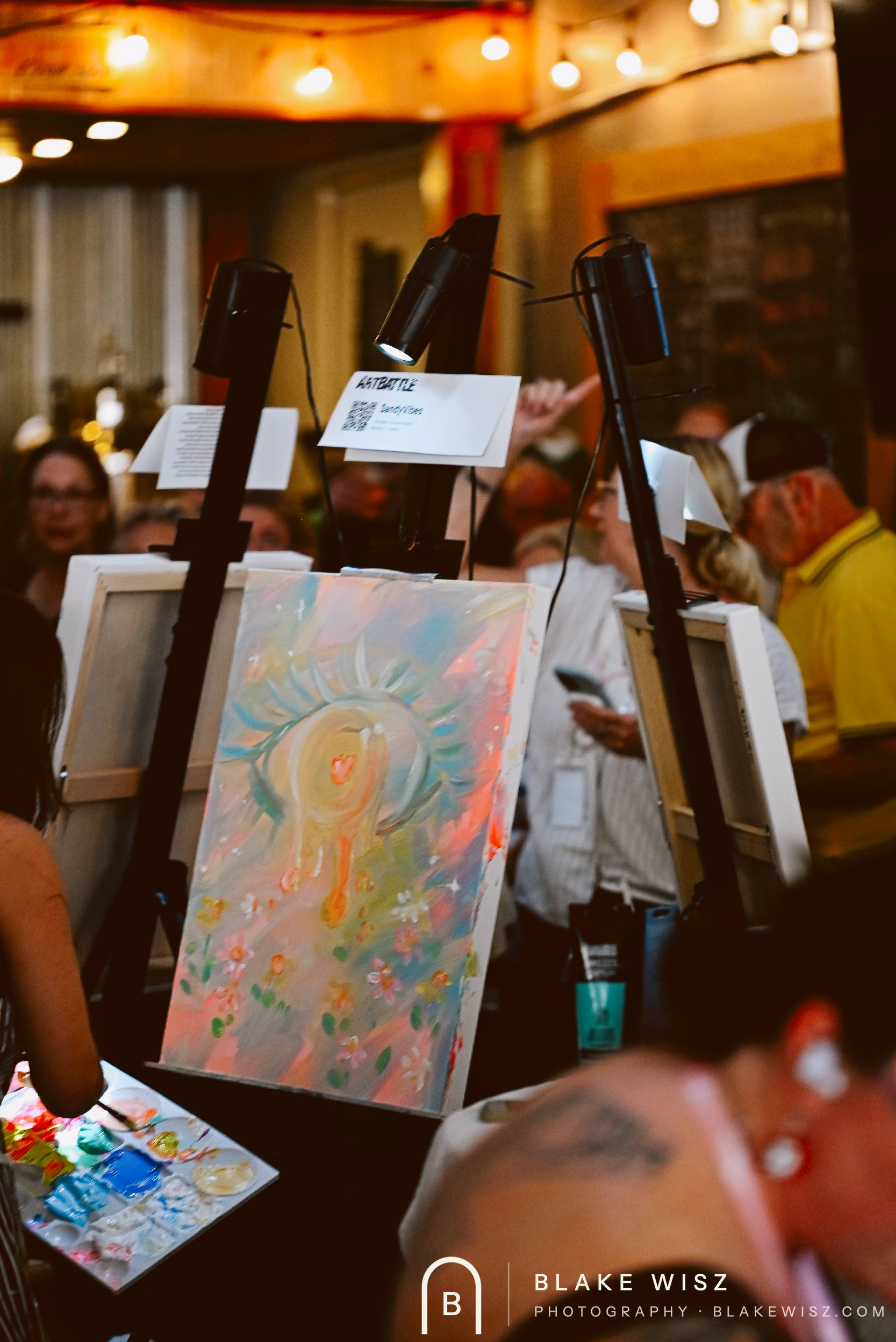 Finished sun and flower canvas lit on its easel beside the Art Battle bid card