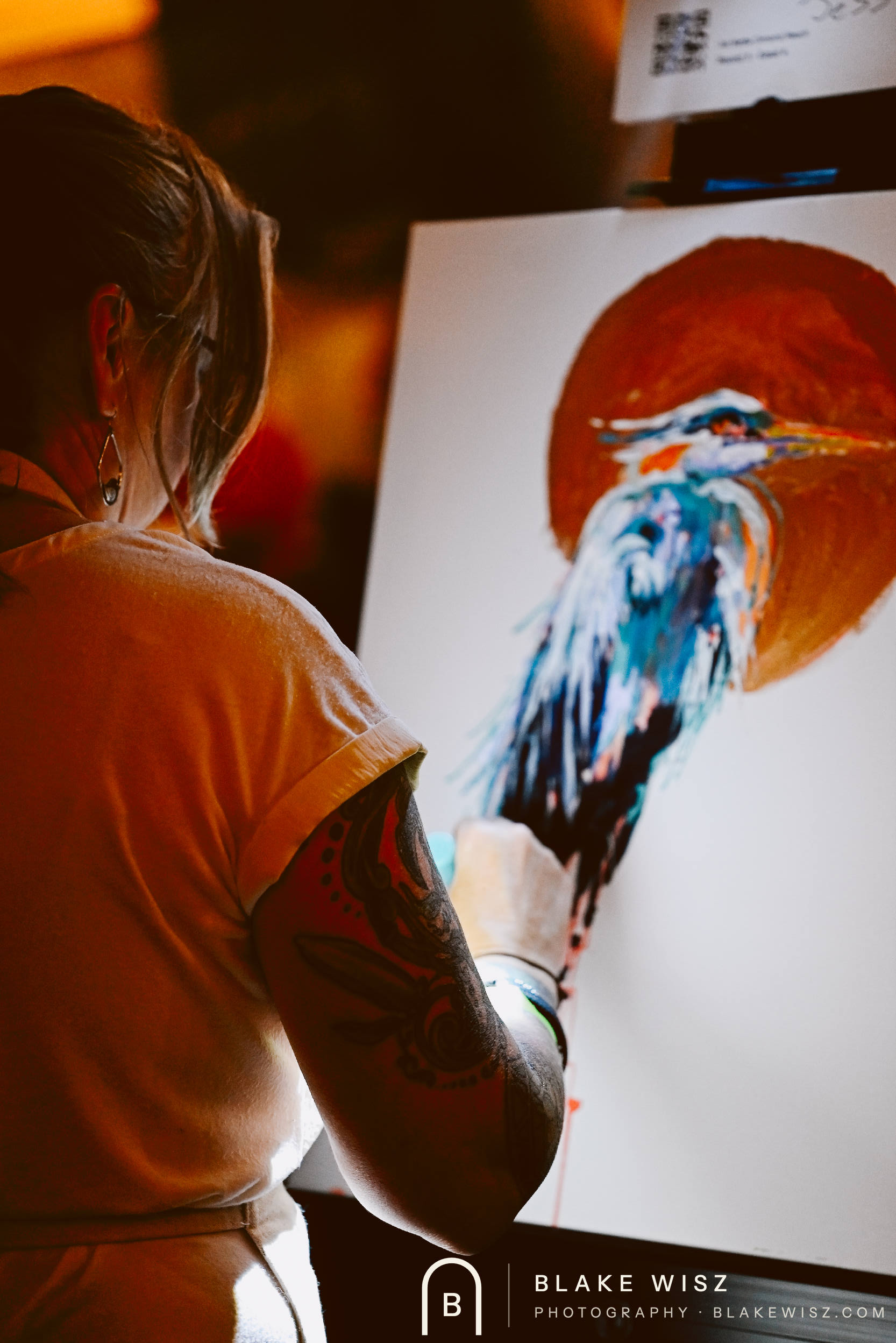 Tattooed artist painting a teal heron against a red sun, photographed from behind