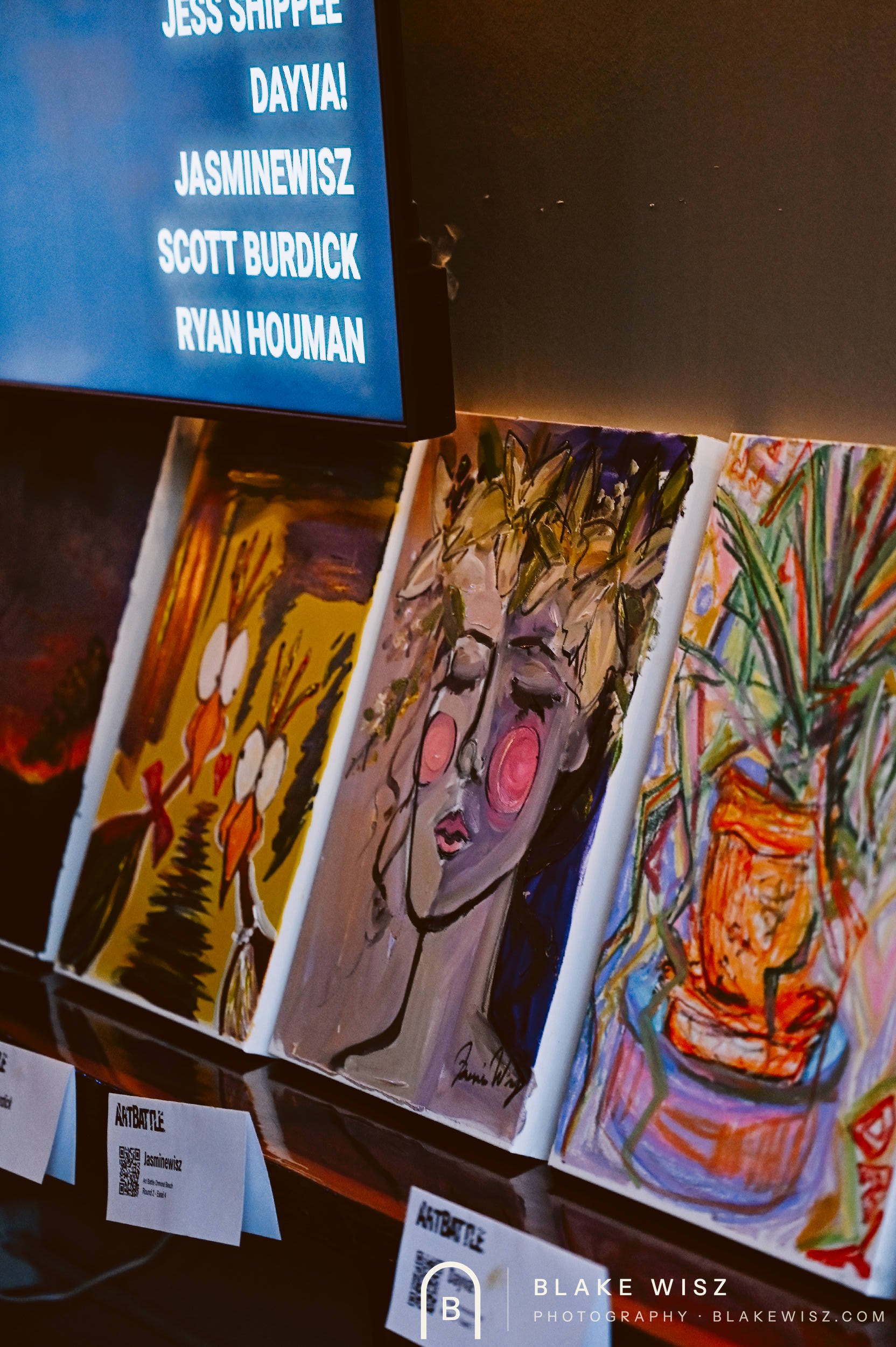 Row of finished Art Battle canvases below a screen listing the competing artists