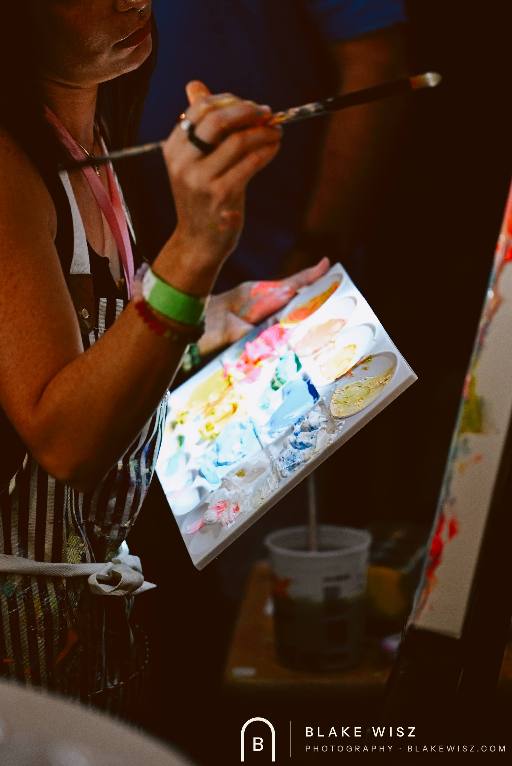 Artist in a striped apron holding a paint-covered palette and brush beside her easel