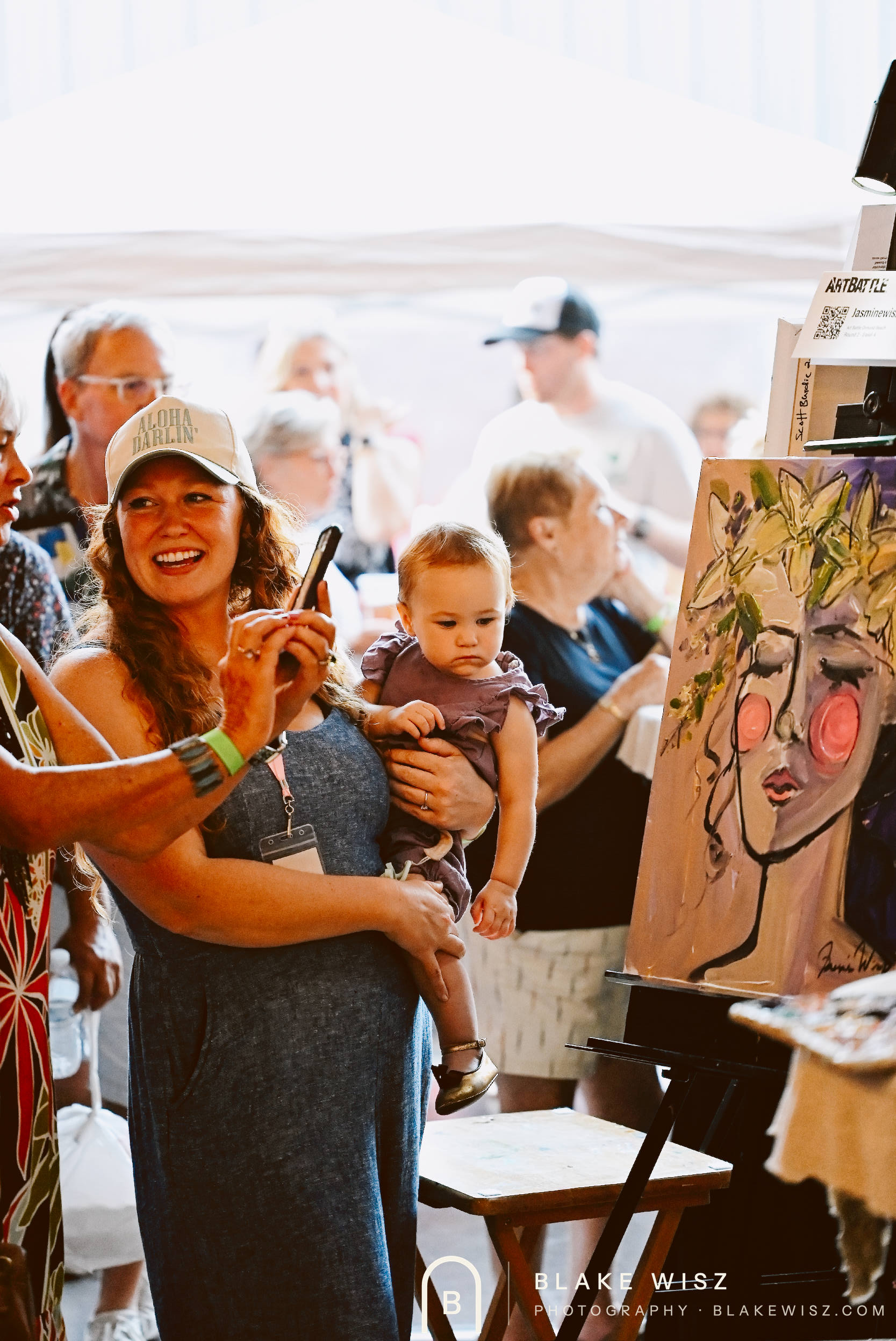Jasmine Wisz photographing her finished Art Battle painting while holding a small child