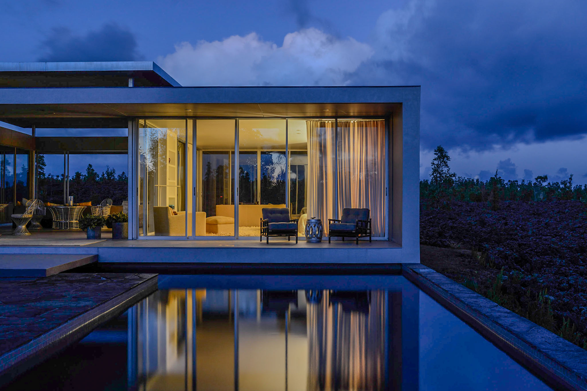The lit glass house reflected in the black pool at dusk