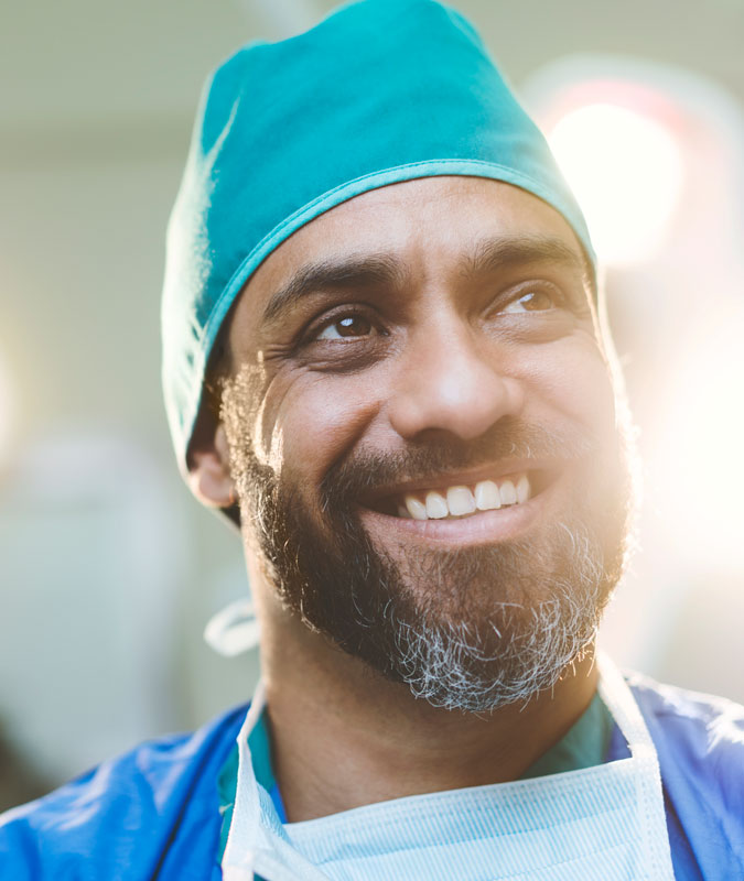 Smiling male surgeon wearing a teal surgical cap and gown with a mask around his neck in a brightly lit operating room.