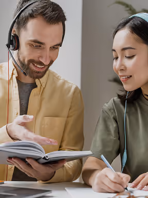 Man wearing a headset with microphone pointing at an open book while a woman writes notes beside him, both seated at a desk.