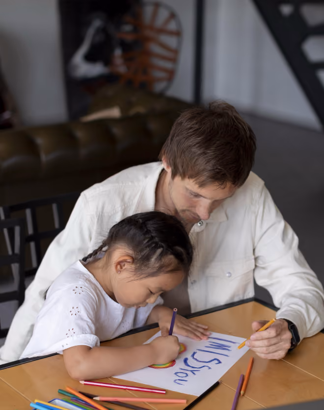 Man and young girl sitting at a table drawing and coloring on a piece of paper with colored pencils, the girl is coloring a rainbow while the man holds a pencil and watches.