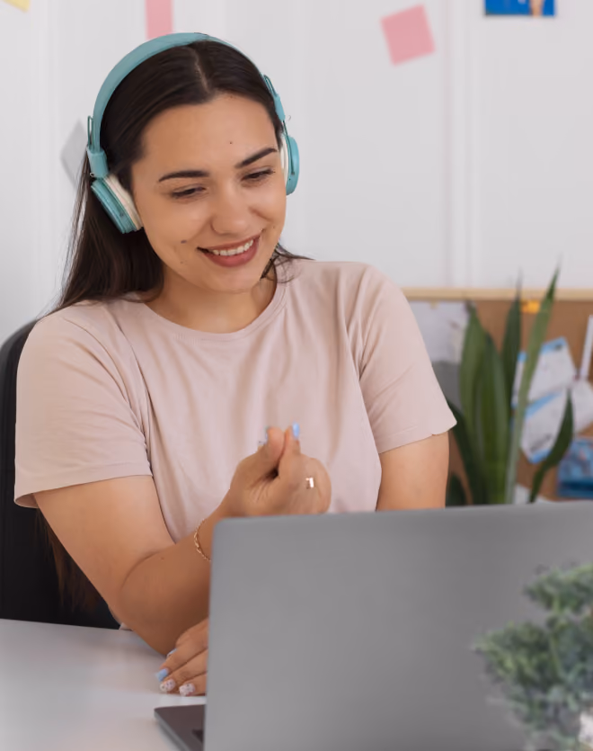 Smiling woman with dark hair wearing mint green headphones and a light pink t-shirt, sitting at a desk and gesturing while looking at a laptop.
