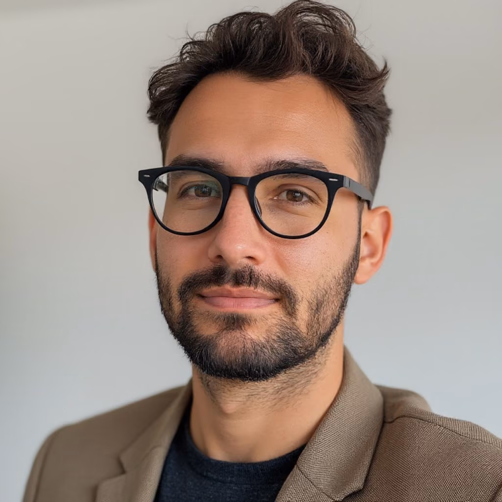 Portrait of a young man with short, dark wavy hair, a beard, and black-rimmed glasses, wearing a brown blazer over a dark shirt, against a plain light background.