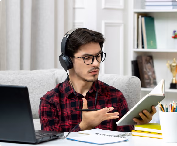 A young man wearing glasses and headphones studies at a desk with a laptop, notebooks, and colored pencils, holding a book open in one hand and gesturing with the other.