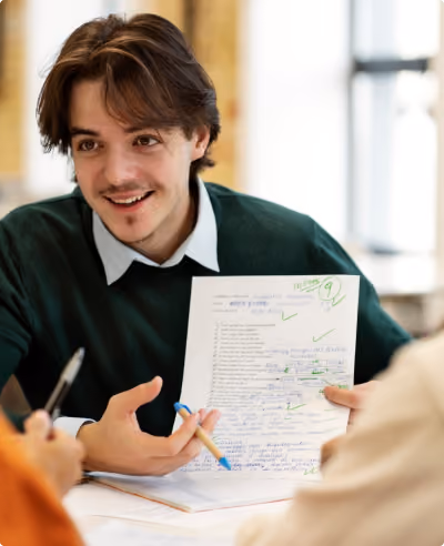 A young man with brown hair and a mustache in a dark green sweater holds a graded paper with green check marks and a score of 91 while talking to two people across a table in a light-filled room.