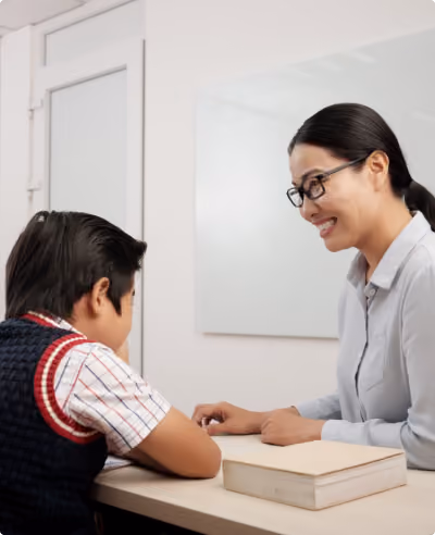A woman wearing glasses and a gray shirt smiling at a young boy sitting across a table with a book on it in a bright room.