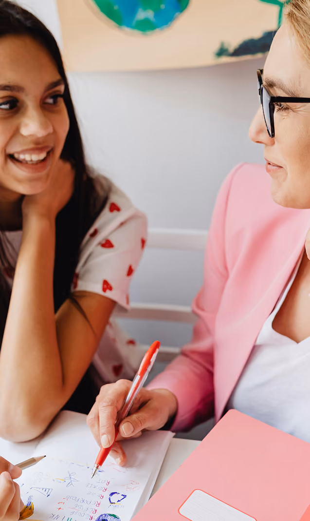 A smiling young girl with long dark hair talks to a woman wearing glasses and a pink blazer who is holding a red pen and a pink folder, looking at a sheet of paper with colorful drawings and notes.