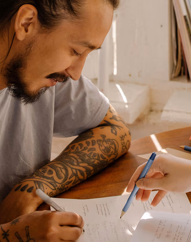 Man with a beard and tattooed arm writing on paper at a wooden table while another person points at the paper with a pen.