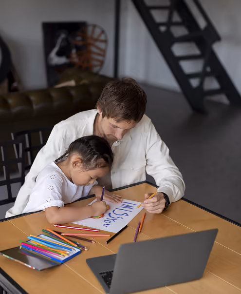 A man and a young girl sitting at a table indoors, drawing with colored pencils on paper. The girl is focused on her drawing while the man helps by holding the paper. Colored pencils and a laptop are on the table.