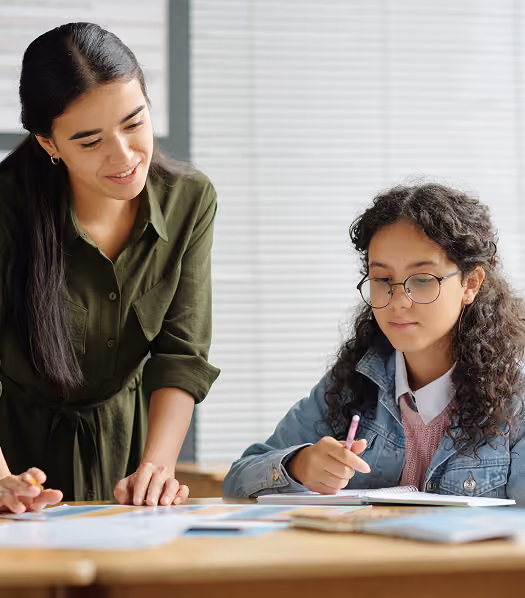A young female teacher with long dark hair and a green shirt smiles while helping a student with curly hair and glasses, who is writing with a pencil at a desk covered in papers.