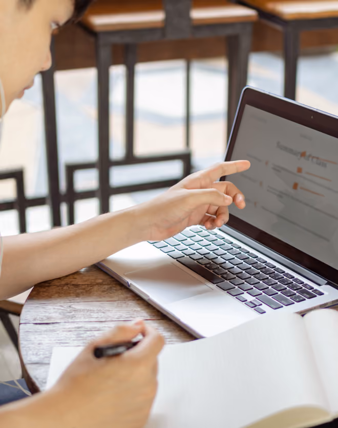 Person pointing at a laptop screen while holding a pen and writing in a notebook at a wooden table, with chairs in the background.