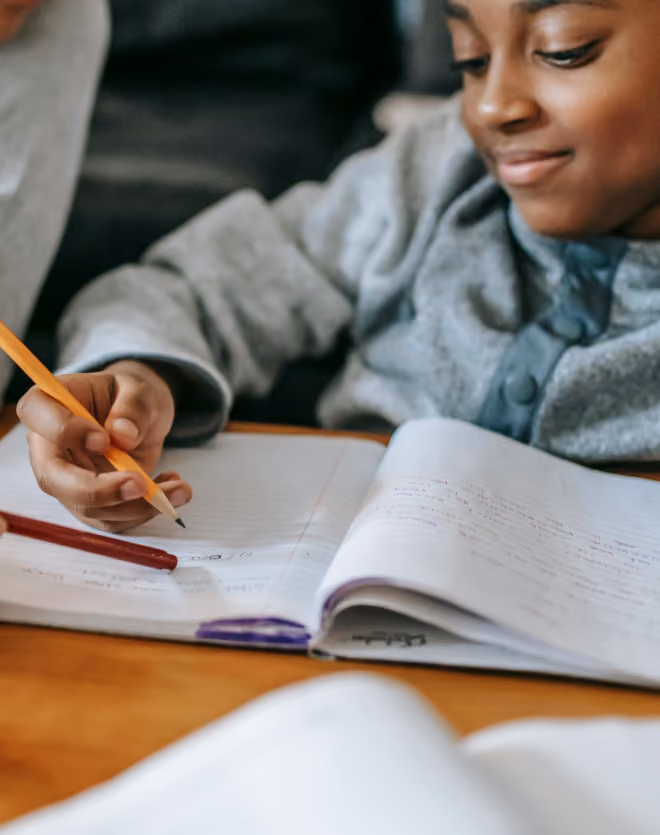 A child wearing a gray sweater is smiling and writing in a notebook with a pencil while sitting at a wooden table.