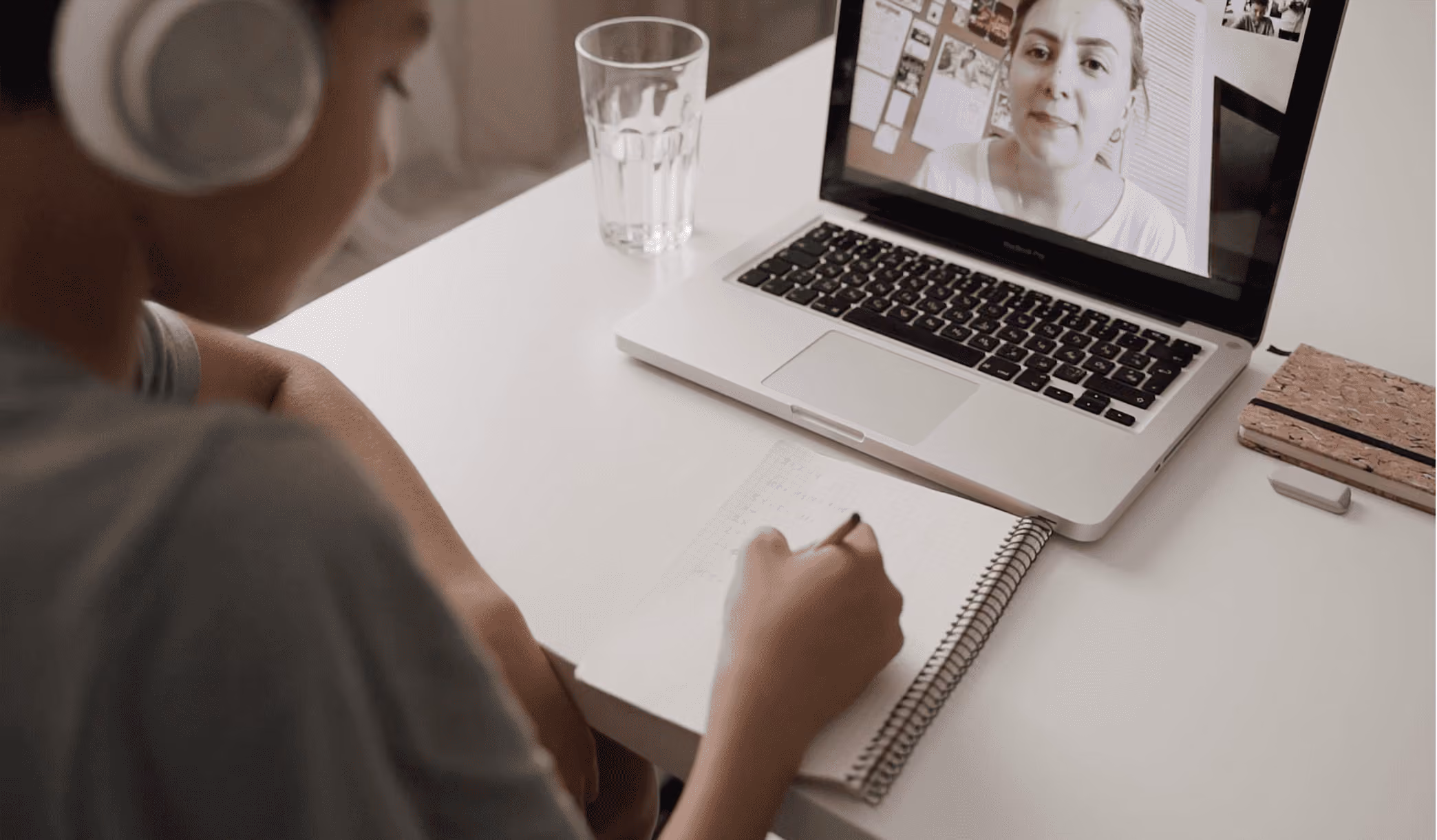 Person wearing headphones writing in a notebook while attending a video call on a laptop with a woman’s face on the screen, sitting at a white desk with a glass of water and notebooks.