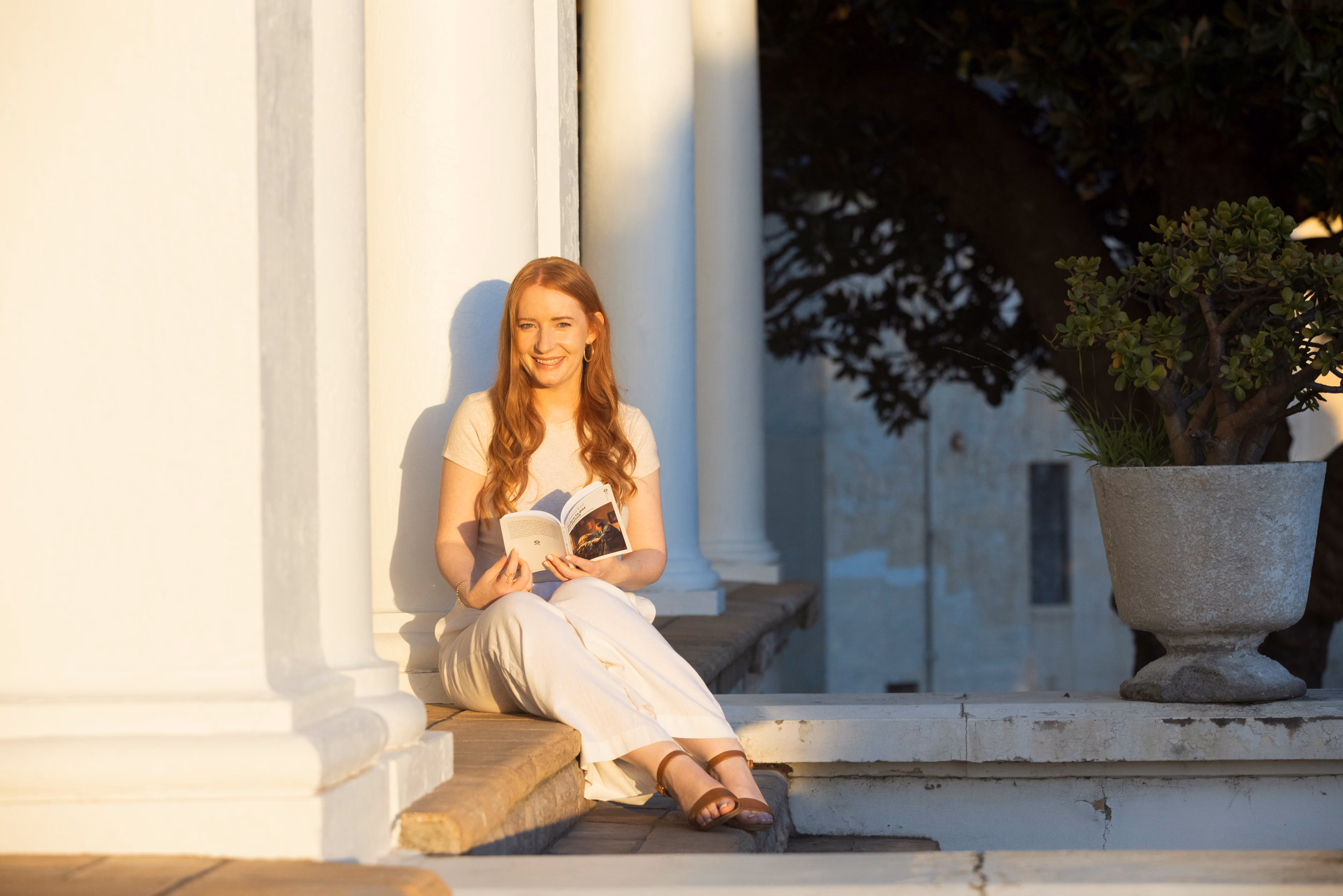 Woman with long reddish hair sitting on outdoor stone steps, reading a book, with white columns and a large potted plant nearby in sunlight.