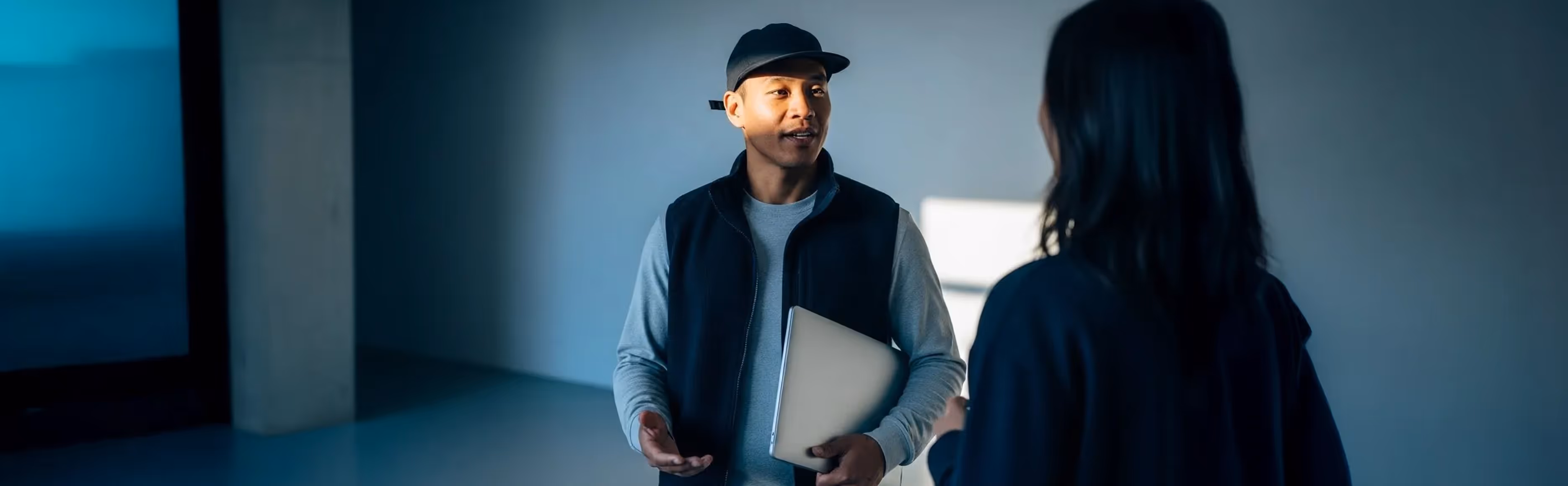 Man wearing a black cap and vest holding a laptop, talking to a woman with long hair in a dimly lit room.