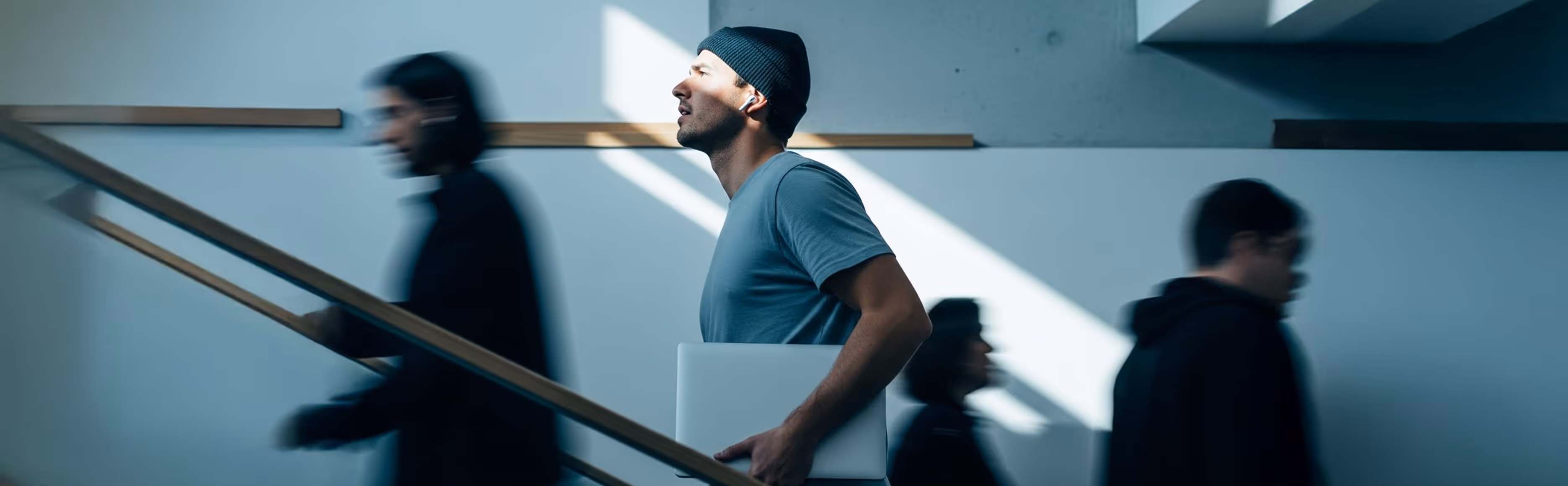 Man wearing a beanie and earbuds carrying a laptop while walking up stairs in a modern building with blurred people around him.