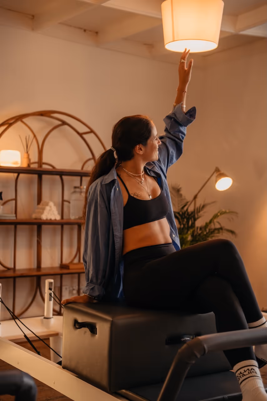 A woman in sportswear and a blue shirt sitting on a pilates reformer machine reaching up to a ceiling light in a warmly lit room.