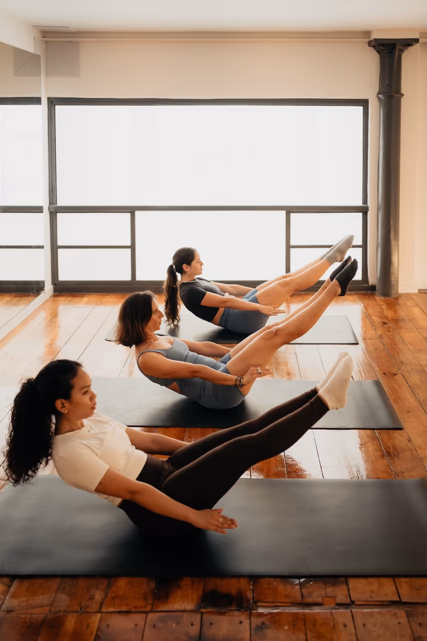 Three women performing a Pilates exercise lying back with legs raised on black mats in a bright studio with wooden floors.