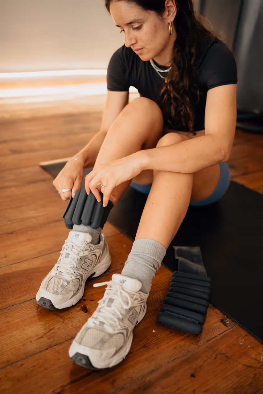 Woman sitting on a yoga mat fastening ankle weights over grey socks and white sneakers on a wooden floor.