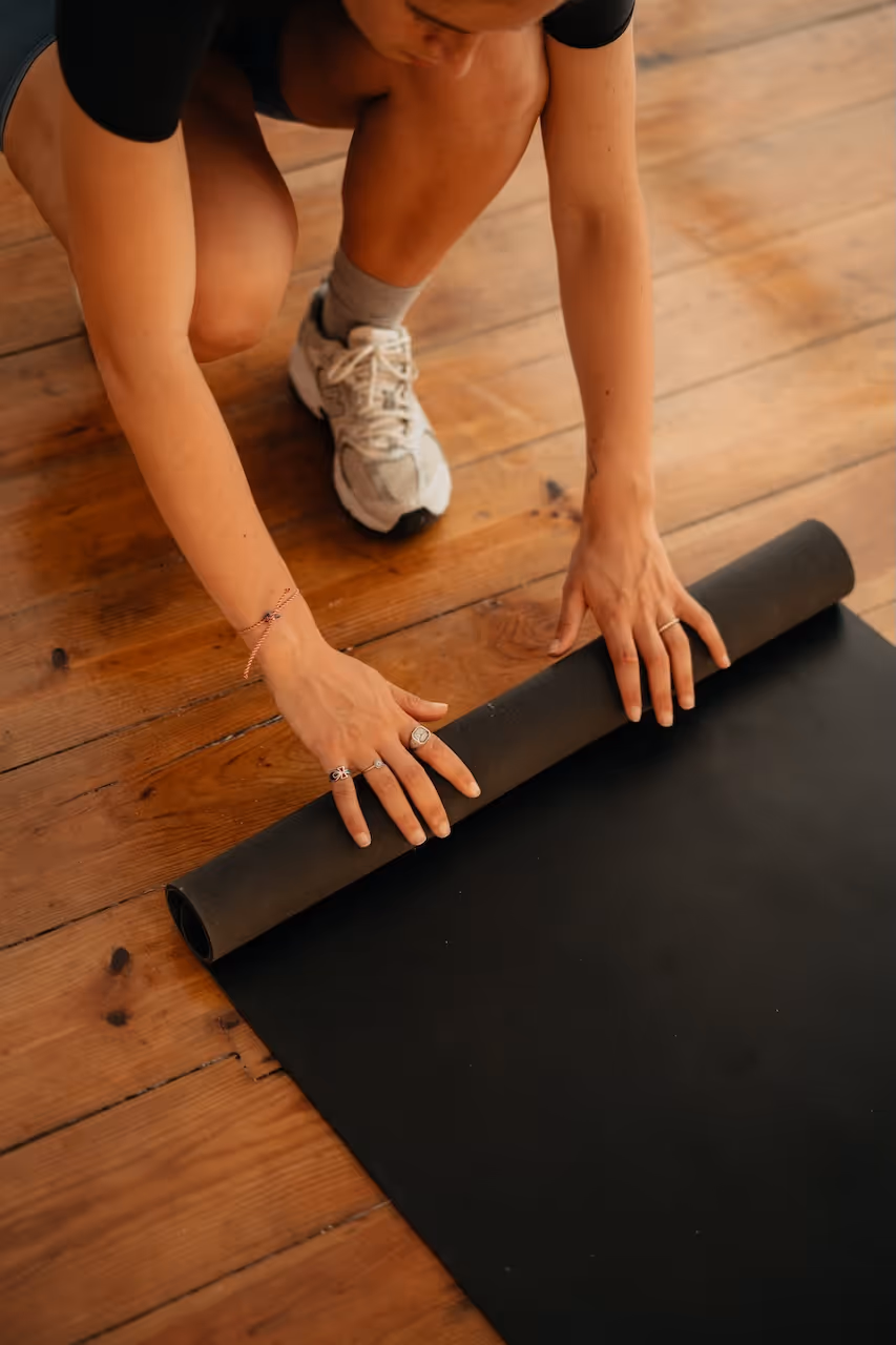Person kneeling on wooden floor rolling out a black yoga mat.