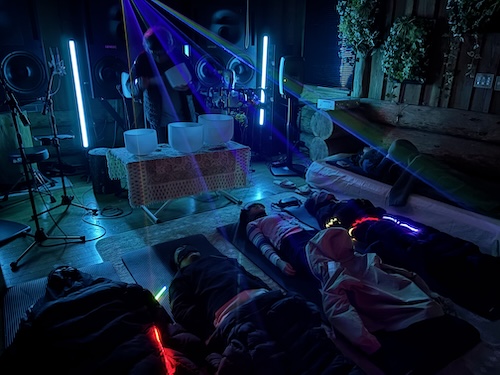 暗い室内で光線が照らす中、マットに寝そべる人々が光るアクセサリーを身につけている。