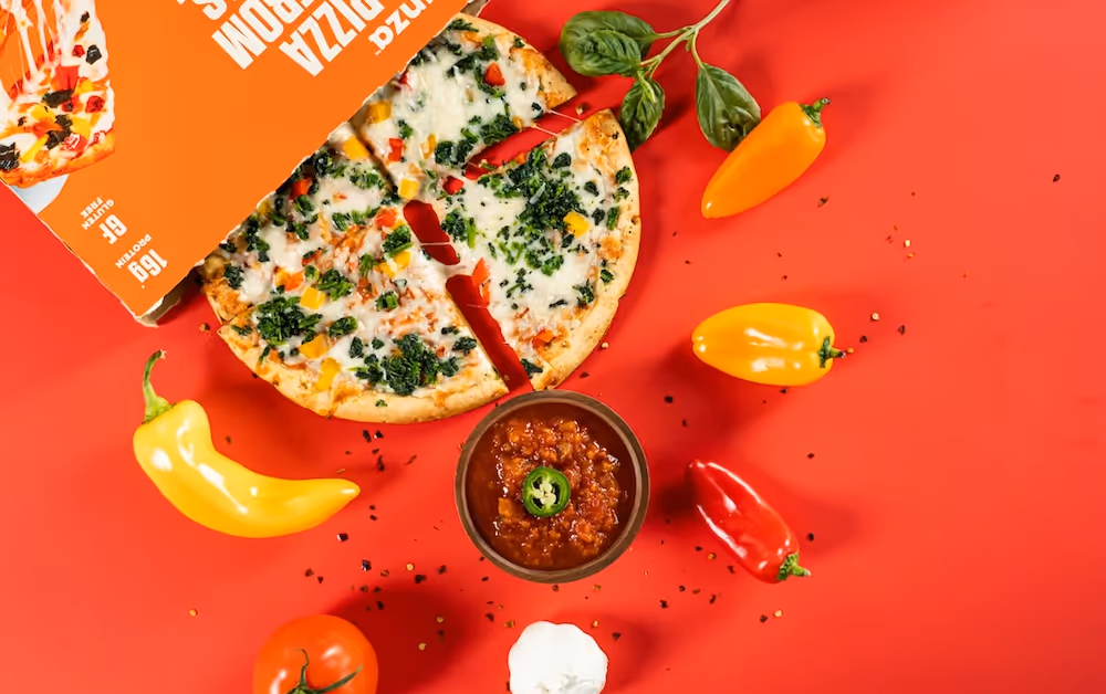 Sliced vegetable pizza with cheese, surrounded by colorful peppers, tomato, garlic, basil, and a bowl of red salsa on a red background.