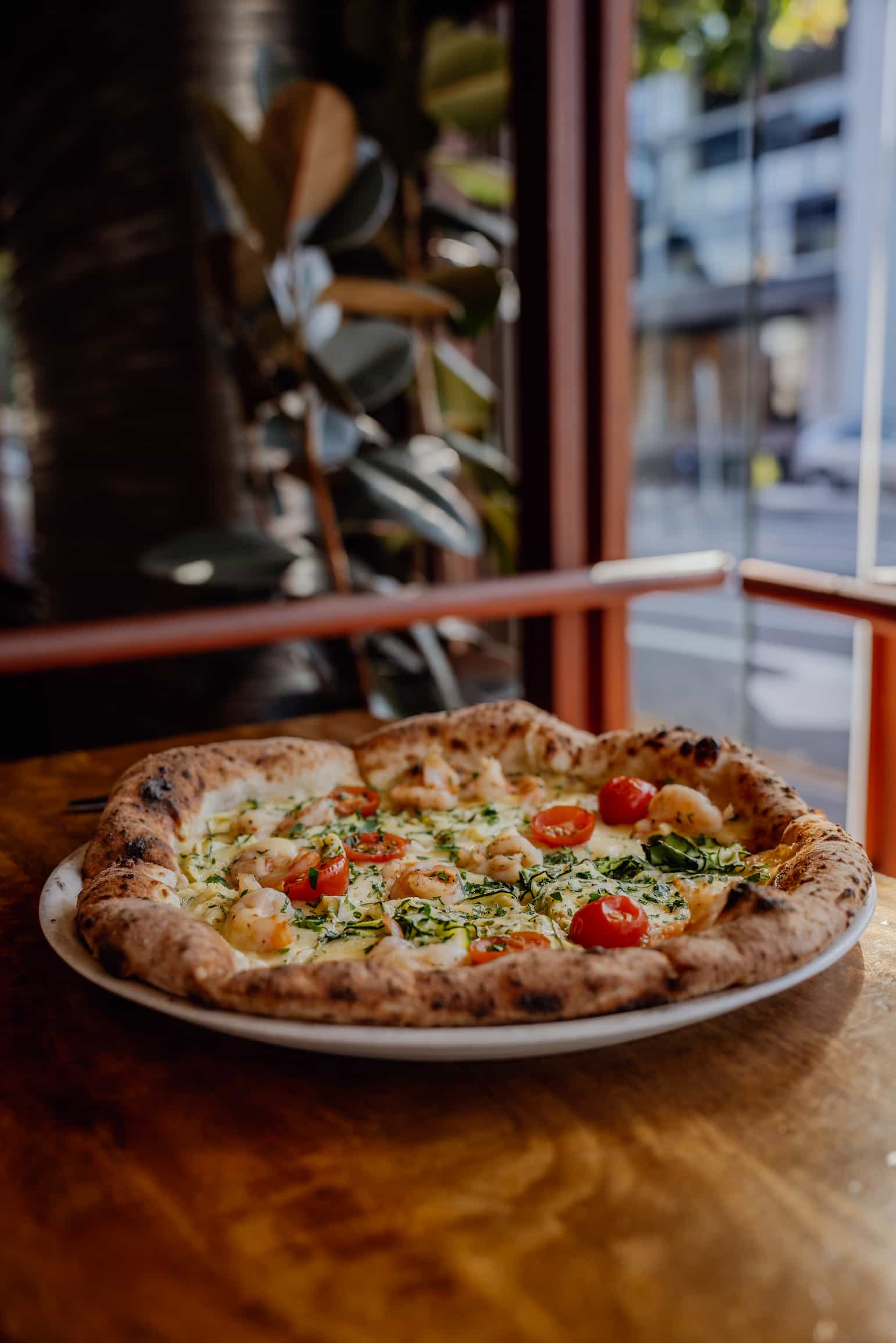 A pizza with cherry tomatoes, cheese, and herbs on a plate rests on a wooden table.