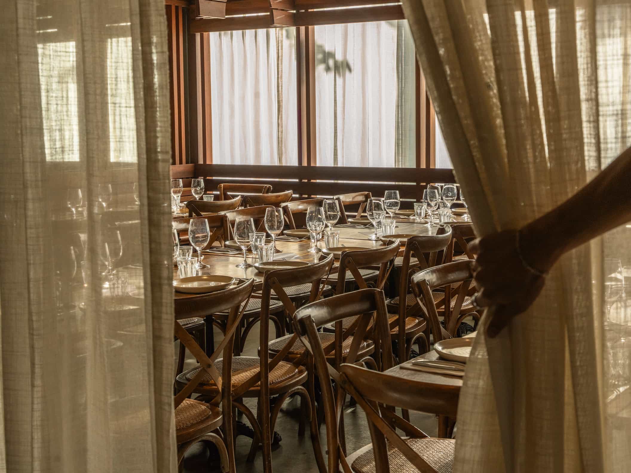 A hand pulls back a curtain to reveal a set dining area with neatly arranged tables and chairs.