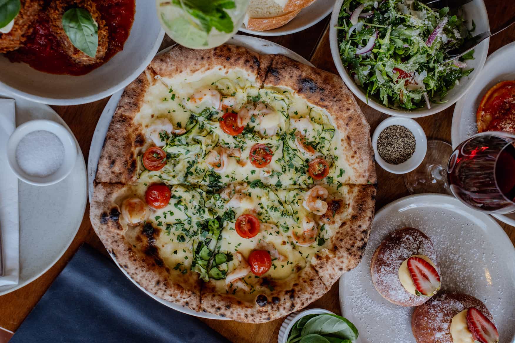 A pizza topped with cherry tomatoes and herbs is surrounded by salads, drinks, and desserts.