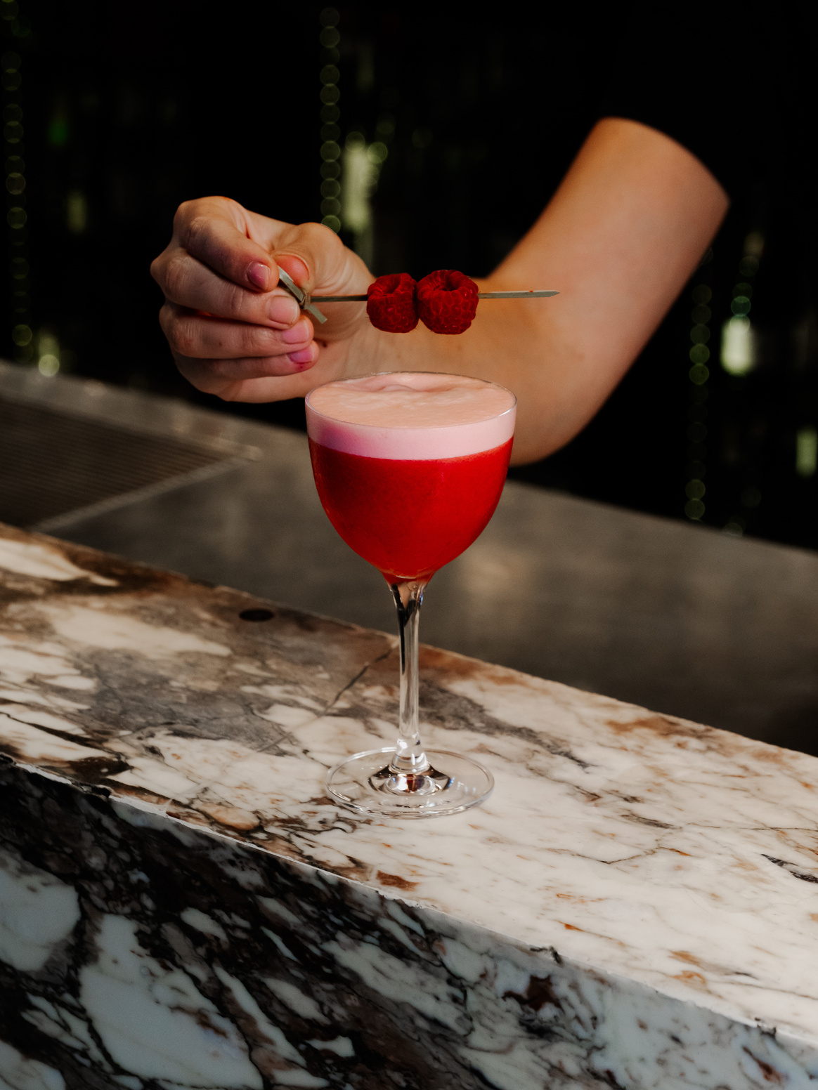 Person holding skewered raspberries above a red cocktail with foam, on a marble countertop.