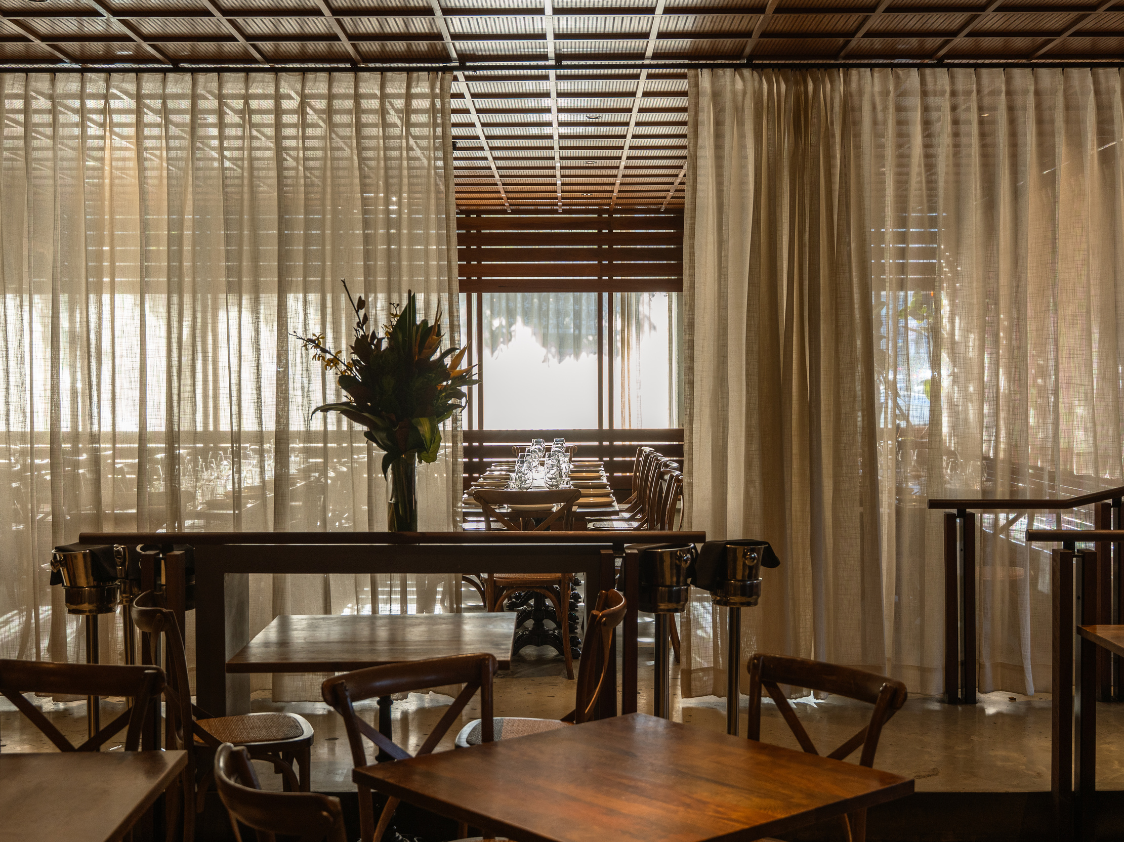 A cozy restaurant with wooden tables and chairs, featuring a floral centerpiece and soft, flowing curtains.
