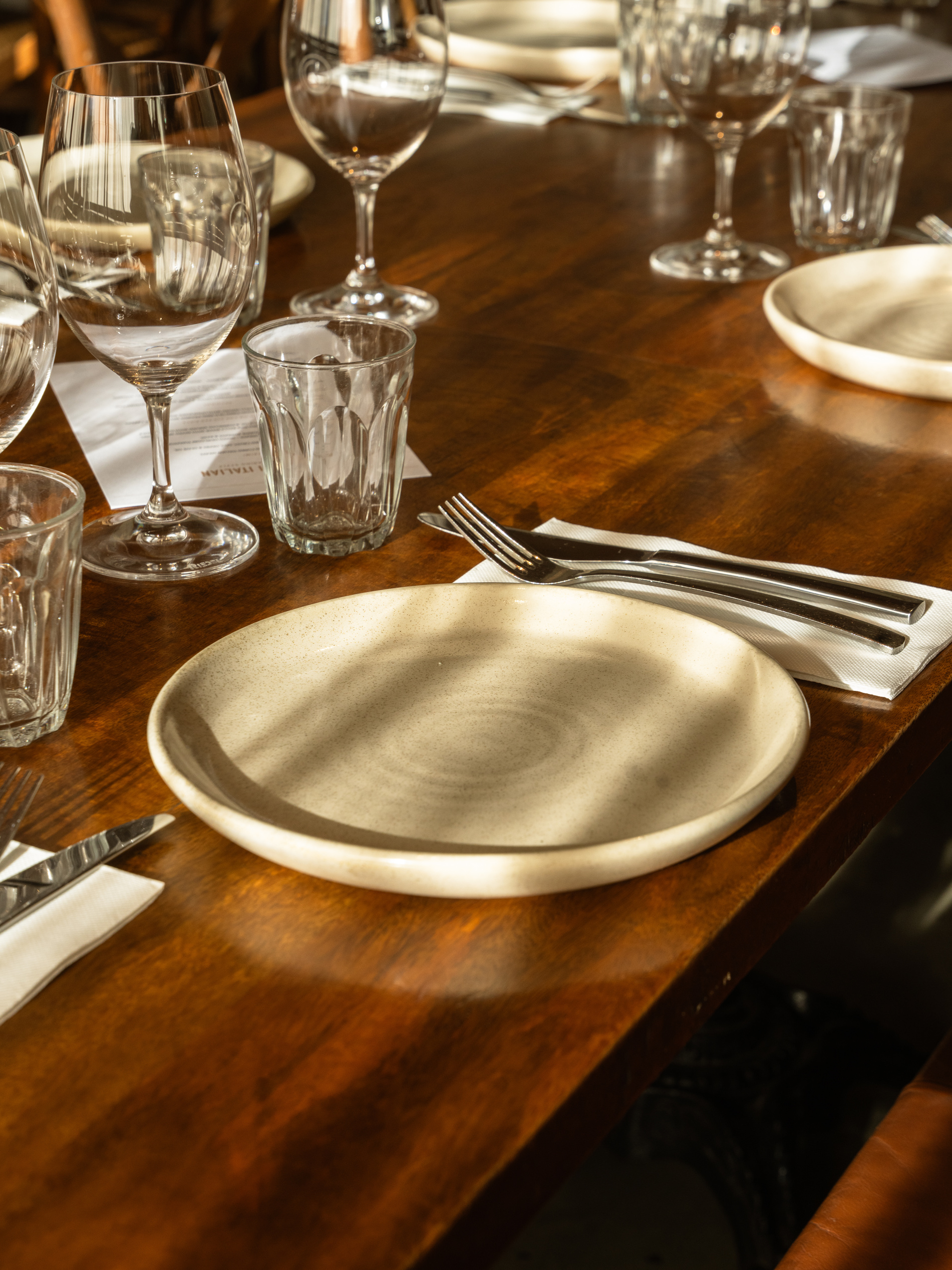 A wooden table set with empty plates, glasses, and silverware in a warm, sunlit setting.