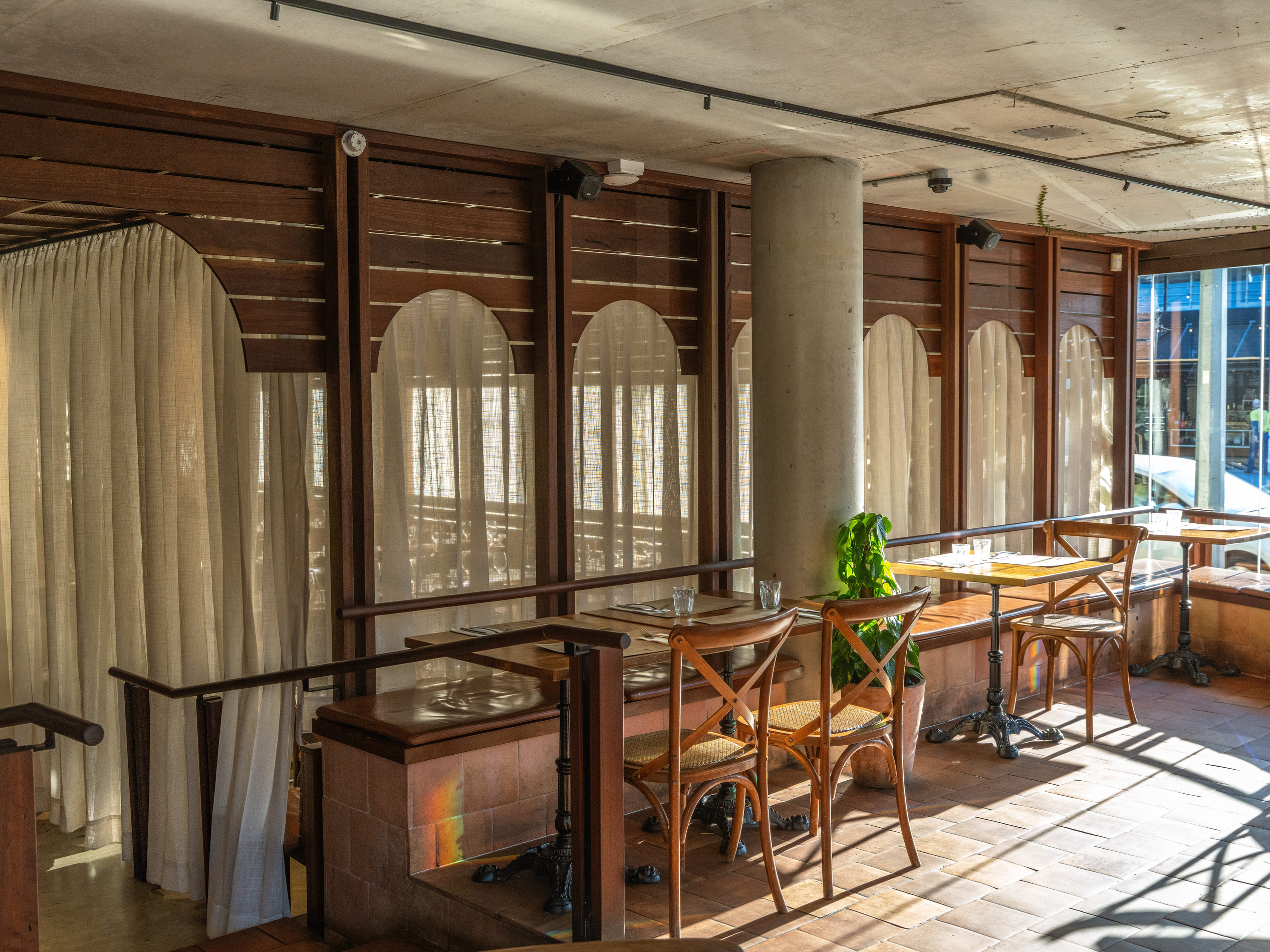 Empty indoor restaurant setting with wooden tables and chairs, lit by sunlight through large windows.