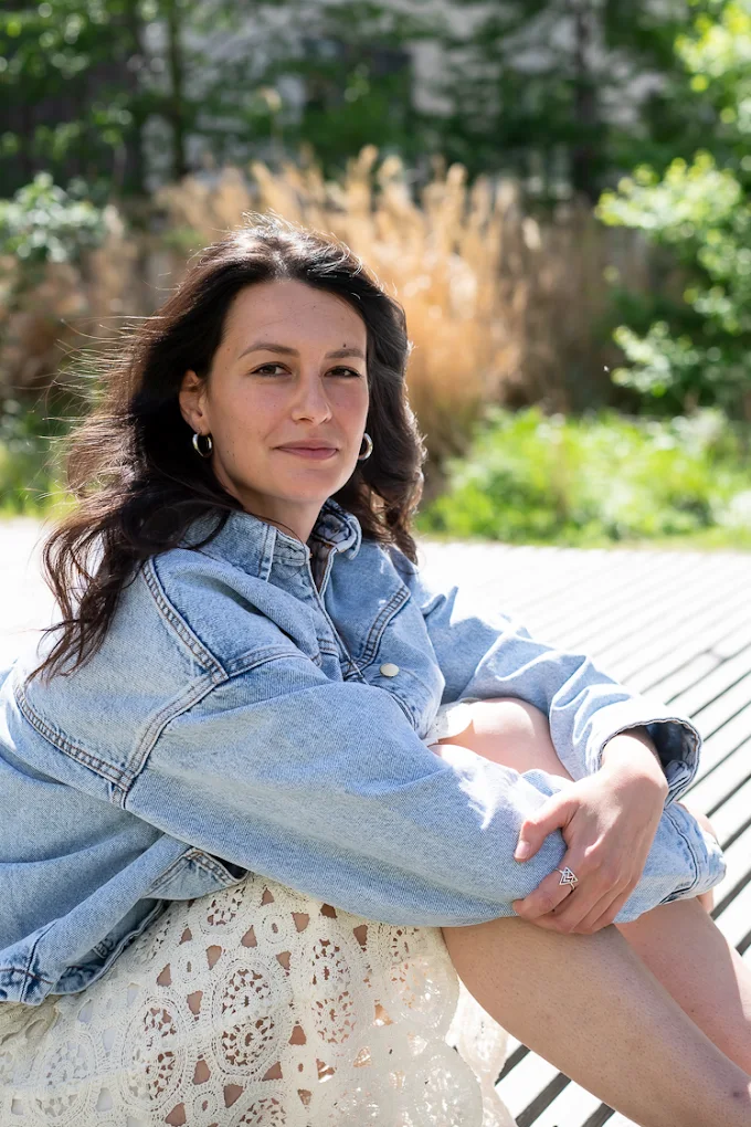 Portrait de Maeva, assise en nature les mains qui tiennent ses jambes repliées mais tournées vers la droite. Le regard vers toi. Une femme douce qui fait de la réflexologie plantaire, des soins énergétiques, du massage.