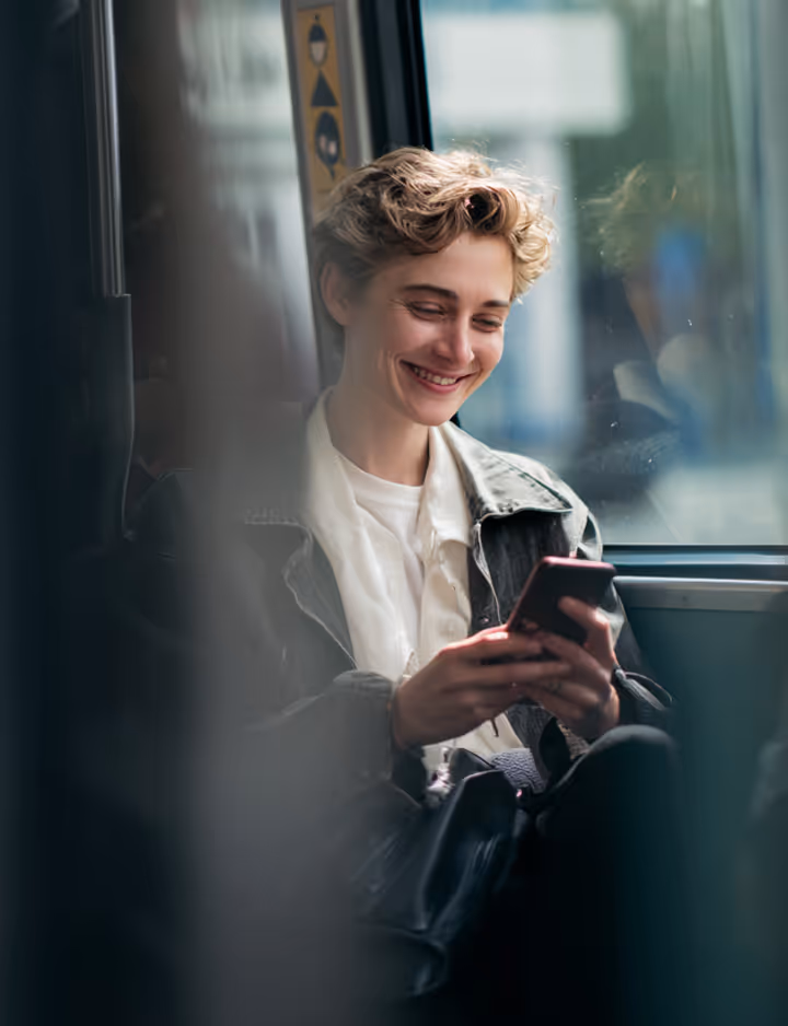 Young person with short curly hair smiling while looking at a smartphone on public transit.