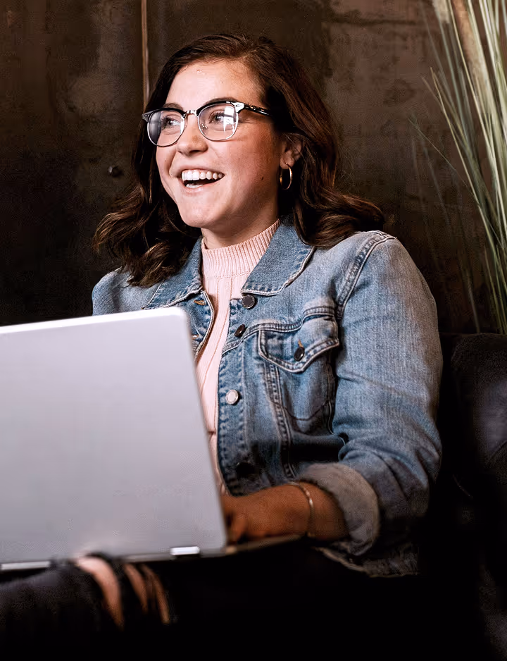 Smiling woman with glasses wearing a denim jacket, working on a laptop.