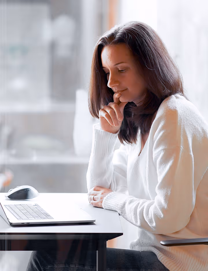 Woman in white sweater sitting at a desk looking thoughtfully at a laptop.