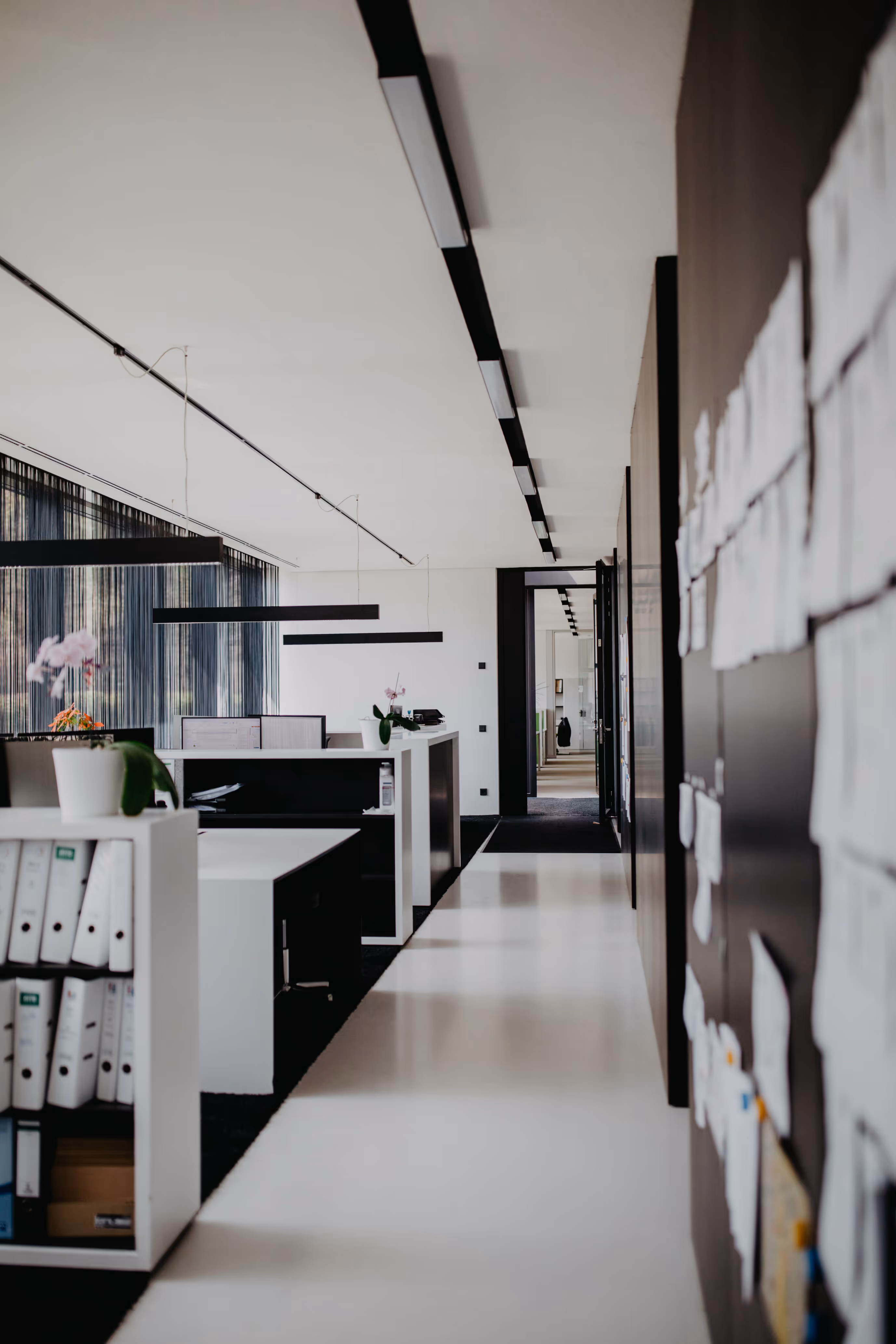 Moderner Büroflur mit weißen Schreibtischen, schwarzen Trennwänden, Aktenordnern und Pinnwand mit vielen Notizen.