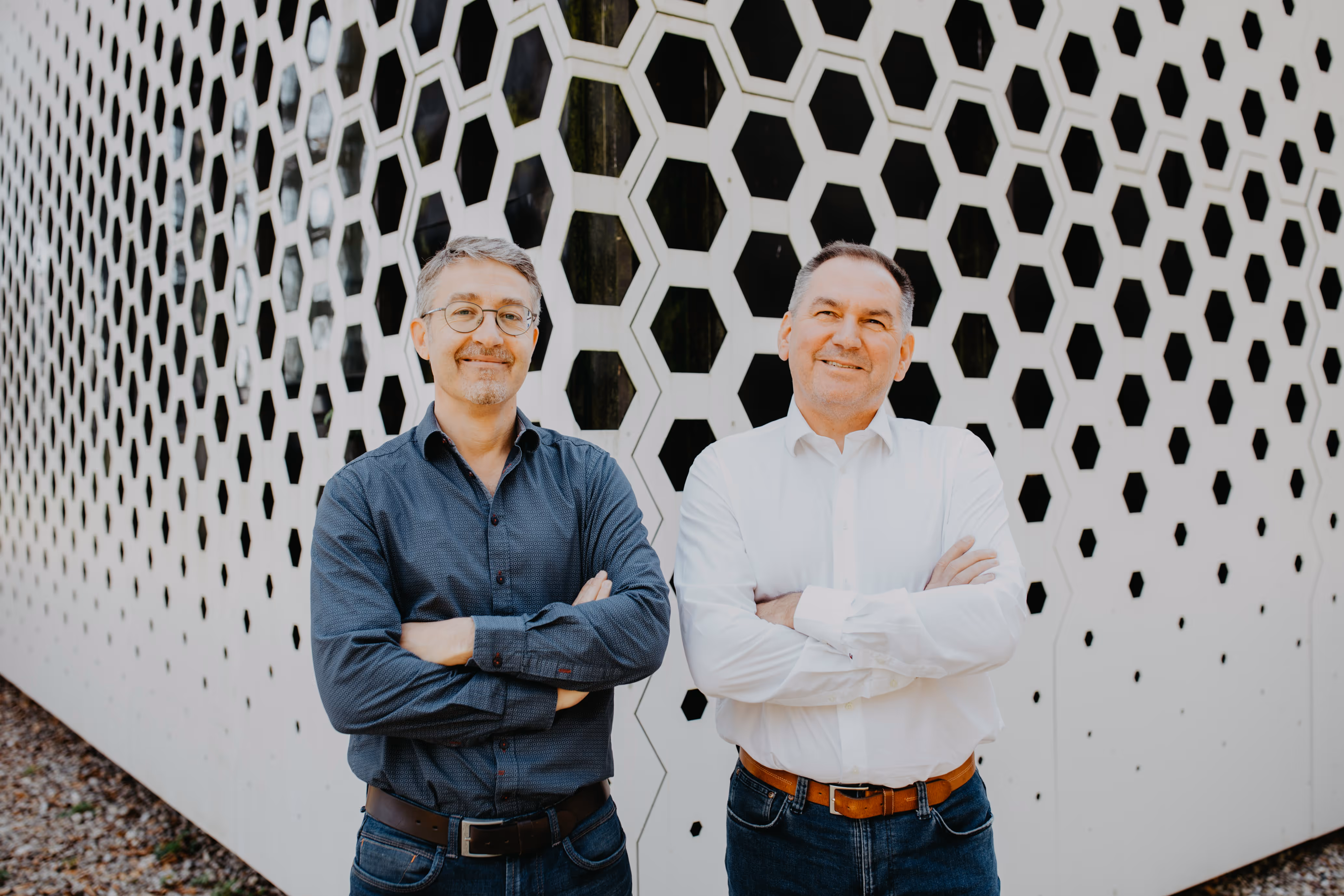 Zwei lächelnde Männer mit verschränkten Armen stehen vor einer Wand mit hexagonalem Muster.