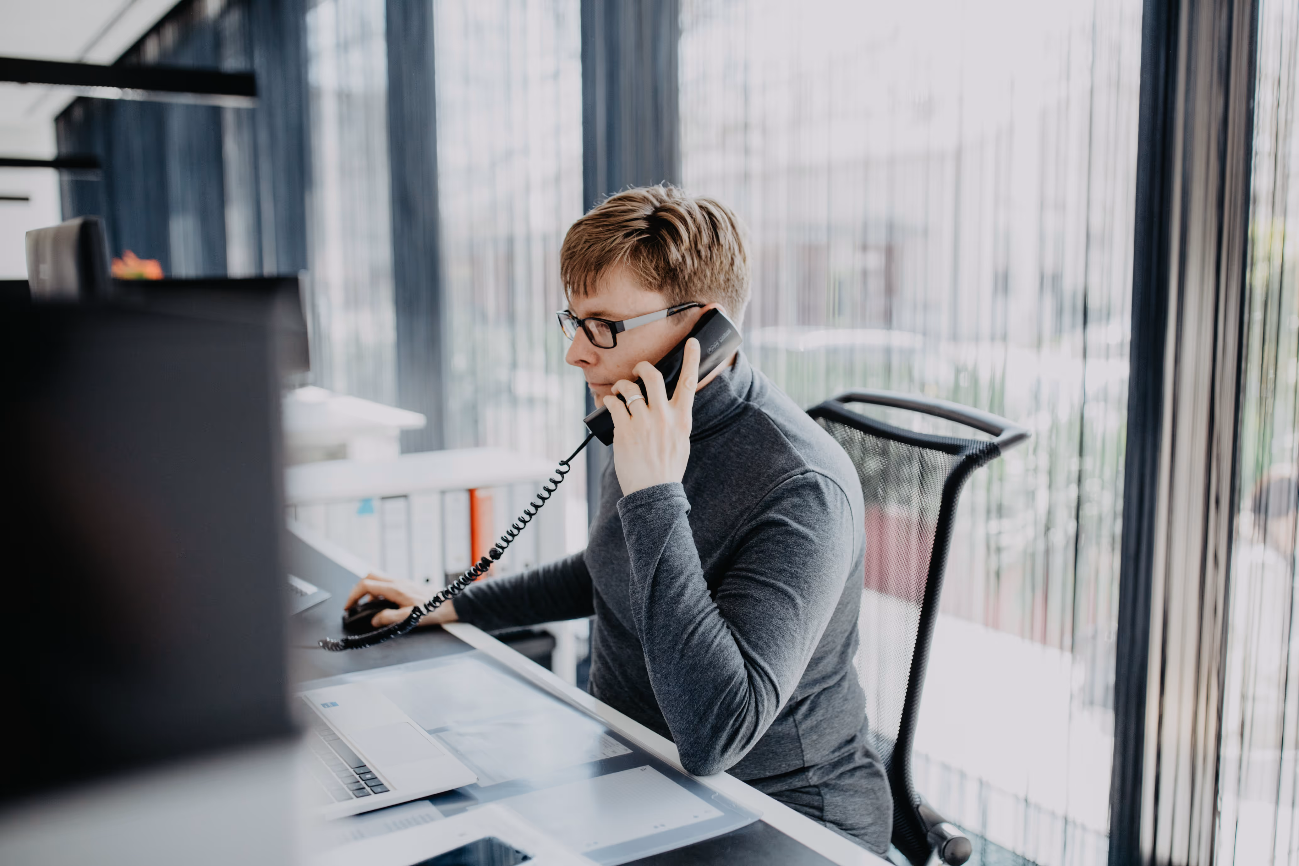 Ein junger Mann mit Brille sitzt an einem Schreibtisch und spricht am Telefon, während er einen Computer bedient.