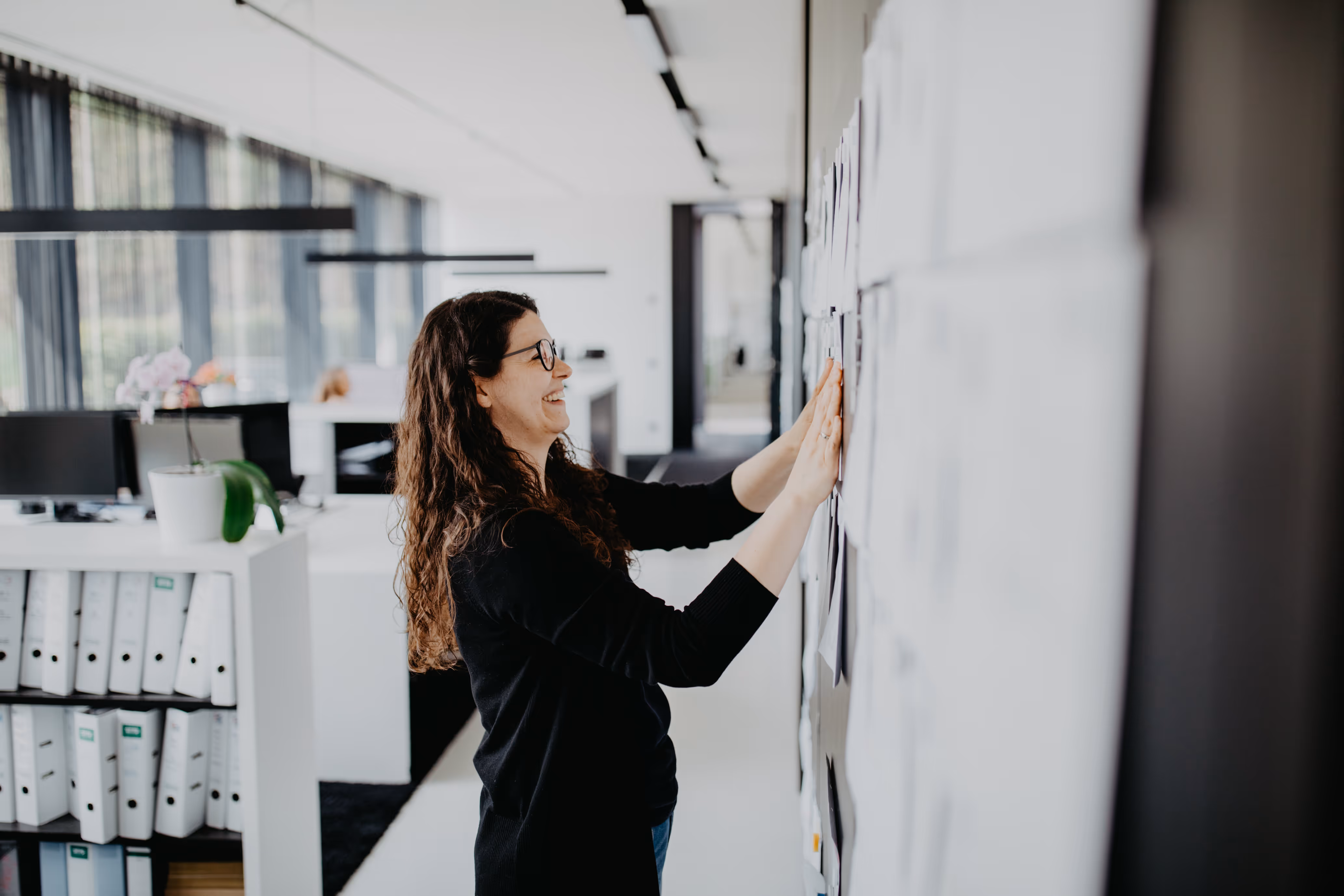 Frau mit langen braunen Haaren und Brille lächelt, während sie an einer mit Notizen bedeckten Wand in einem modernen Büro arbeitet.