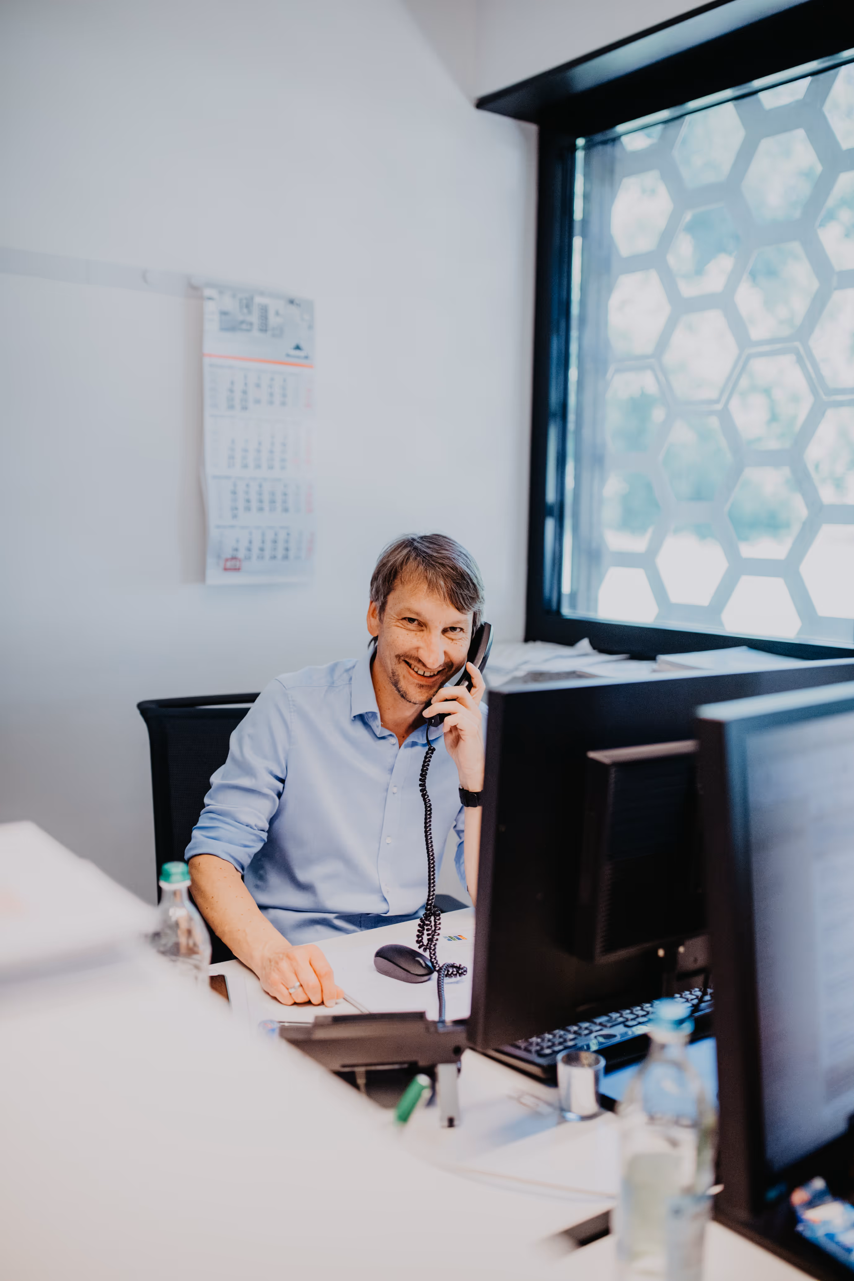 Mann im Büro sitzt am Schreibtisch und telefoniert lächelnd, vor ihm zwei Computermonitore und eine Fensterwand mit Wabenmuster.