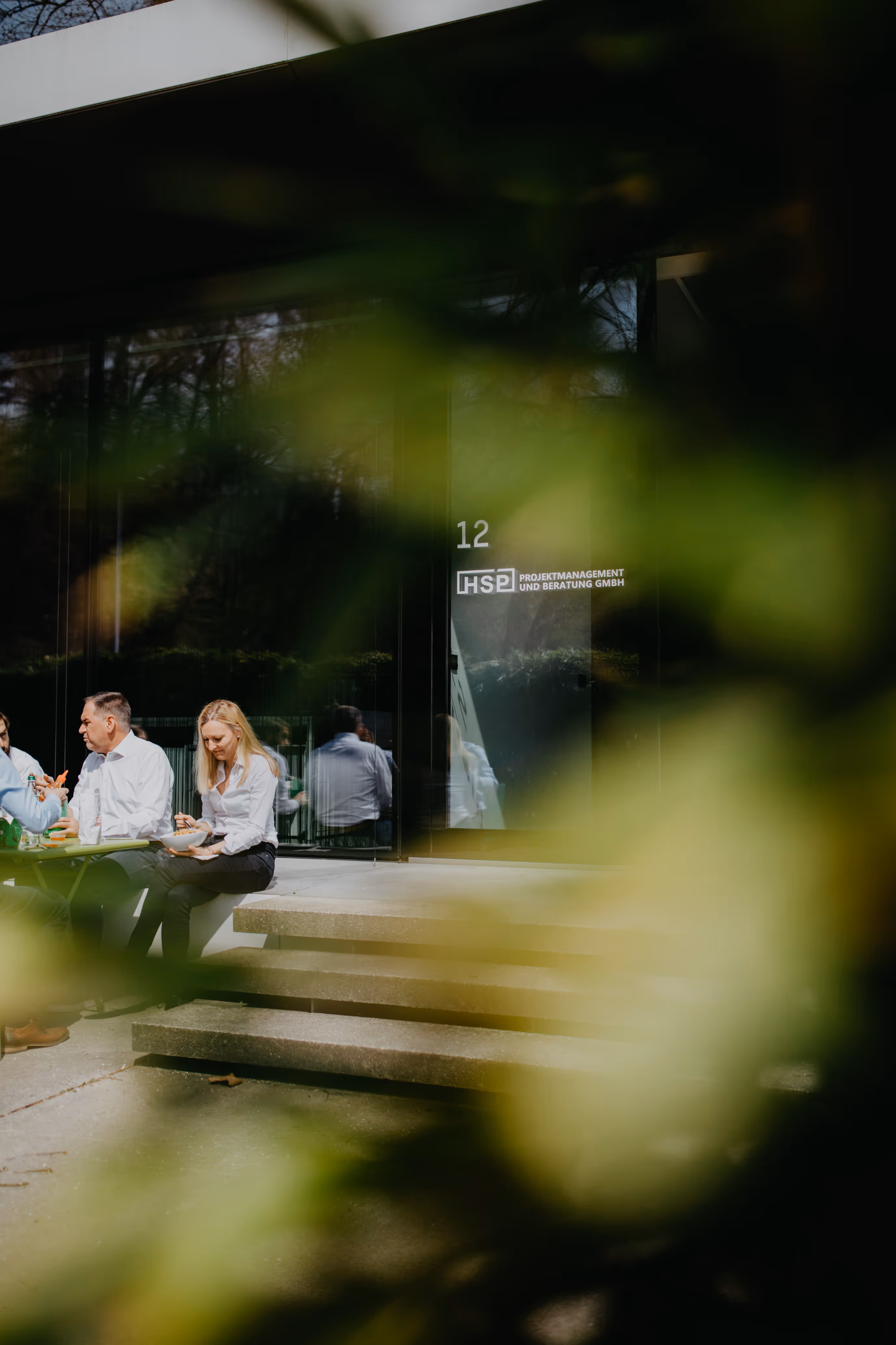 Drei Personen sitzen draußen vor einem modernen Gebäude mit Glasscheiben und essen zusammen bei Sonnenlicht.